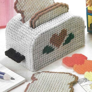 May include: A white crocheted toaster with a brown and green heart design. The toaster has two slices of bread popping up. There are also several crocheted bread slices and a few orange and yellow crocheted circles.