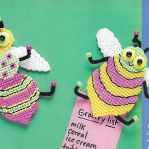 May include: Two handmade, colorful bee-shaped crafts. The bees have yellow, pink, and green bodies with white wings. They have button eyes and pipe cleaner antennae and legs. One bee is attached to a pink grocery list.