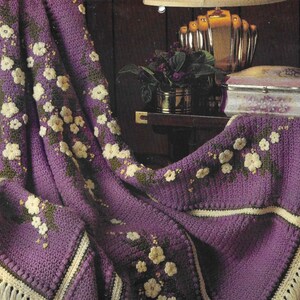 May include: A purple crocheted throw blanket with a white floral pattern and a fringe border. The blanket is draped over a chair and a table with a decorative box.