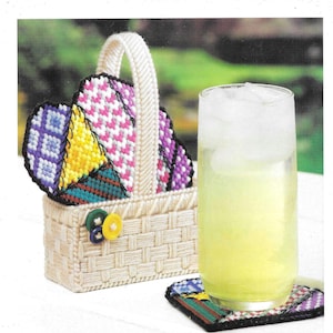 May include: A white wicker basket with a handle holds colorful heart-shaped coasters. The coasters are made of plastic canvas and feature a patchwork design with purple, pink, yellow, and green squares. A glass of lemonade sits on a coaster.