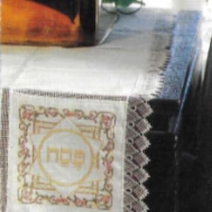 May include: A white table runner with a lace border and a small embroidered design featuring the Hebrew word "Shabbat" in gold. The table runner is on a black table with a woven pattern.