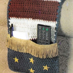Op de afbeelding: Gehaakte armleuningshoes met een rood-wit-blauw gestreept ontwerp en een blauw gedeelte met gouden sterren. De hoes heeft een zak met een franje rand en een afstandsbediening zit in de zak.