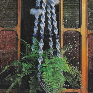 May include: A blue macrame plant hanger with a tassel, hanging in front of a stained glass window. The plant hanger is decorated with blue beads and has a fern plant hanging from it. The text "BLUE BIRD OF HAPPINESS" and "Pattern on Page 13" is visible below the plant hanger.