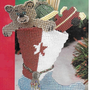 May include: A blue, red, and white stocking with a brown teddy bear, a green bag, and a red toy truck sticking out of the top. The stocking has a gold belt with a silver charm and a beaded tassel.