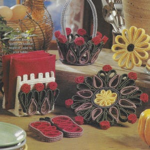 May include: A set of plastic canvas crafts featuring a red and green floral design. The set includes a napkin holder, a small basket, a coaster, and two butterfly-shaped coasters. The text "These fanciful accents bring a splash of festive tropical color to your table." is included in the image.