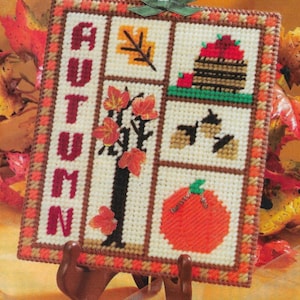 May include: A white canvas with a brown border and a colorful autumn-themed design. The design includes a tree with red and orange leaves, a basket of apples, a pumpkin, and acorns. The word "AUTUMN" is spelled vertically in red and white letters on the left side of the canvas.
