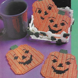 May include: Four orange crocheted pumpkin coasters with black smiling faces. The coasters are in a white crocheted basket with green leaves.