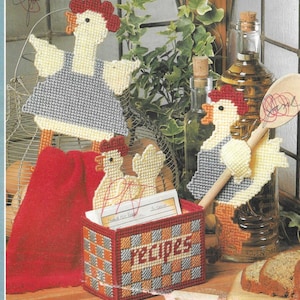 May include: Three crocheted chicken figures with red, yellow and grey accents. The chickens are holding a wooden spoon, a red towel and a recipe box. The recipe box is checkered with orange and grey squares and has the word "RECIPES" printed on the front.