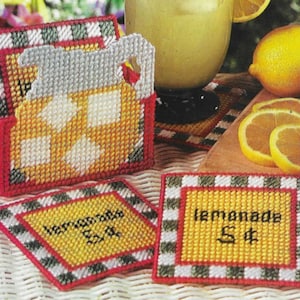 May include: Set of four yellow and white crocheted coasters with black and red checkered borders. The coasters feature the words "lemonade 5¢" and "lemonade cold 5¢". A crocheted pitcher with a yellow and white design is in the background.