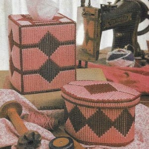 May include: Two pink and brown plastic canvas tissue box covers with a diamond pattern. The tissue box covers are sitting on a brown surface.