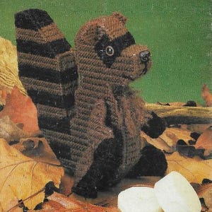 May include: A brown and black crocheted raccoon toy named "Bandit the Raccoon". The toy is sitting on a bed of brown and orange leaves with two white round objects in front of it.