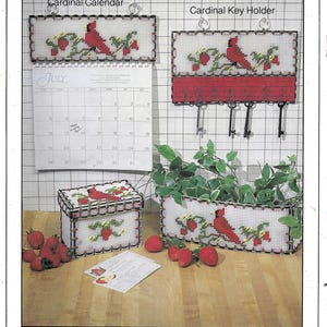 Peut inclure: Un ensemble de quatre organiseurs en point d'aiguille représentant un cardinal avec des accents rouges, blancs et verts. Les organiseurs comprennent un calendrier, un porte-clés, une boîte à recettes et une jardinière. Le calendrier porte l'inscription "Juillet".
