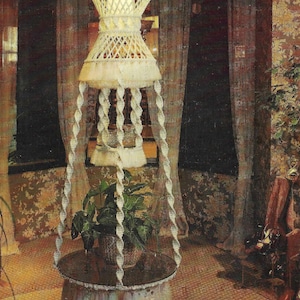 以下が含まれることがあります： 白いマクラメのテーブルの上に植物が置かれています。テーブルは天井から吊り下げられており、その上に白いマクラメのランプシェードがあります。画像には「CRISS CROSS TABLE Pattern on Page 6」というテキストが表示されています。