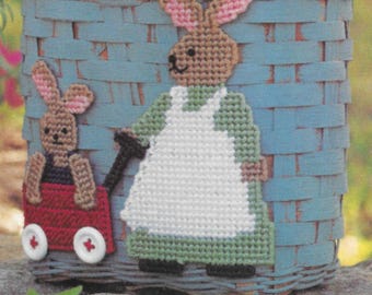 May include: A light blue woven basket with a decorative needlepoint design featuring two bunnies. One bunny is pulling a red wagon, and the other is wearing an apron. The basket is a charming Easter or spring-themed decoration.