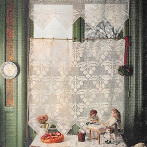 May include: A pair of white crocheted curtains with a delicate floral pattern hang in a window. The curtains are tied back with red ribbon. Two dolls sit at a table in front of the window, with a basket of fruit and a loaf of bread on the table.