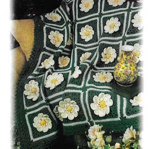 May include: A handmade, green crochet blanket with white and yellow flower appliqués. The blanket is draped over a sofa, showcasing the intricate floral design. A decorative vase with floral patterns sits nearby.