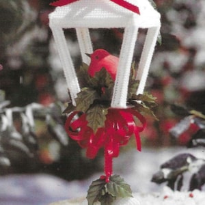 May include: A white plastic lantern ornament with a red ribbon and a red bird inside. The lantern is decorated with green leaves and red berries.