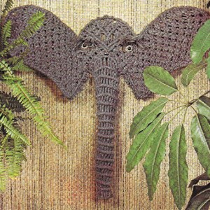 May include: A crocheted elephant head wall hanging in gray with a textured pattern. The elephant is mounted on a woven background with green leafy plants on either side.