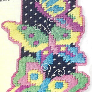May include: A colorful, cross-stitched butterfly design on a dark blue background with white polka dots. The butterfly features sections of yellow, green, pink, and blue. The edges are outlined in pink, and white wire accents are included.