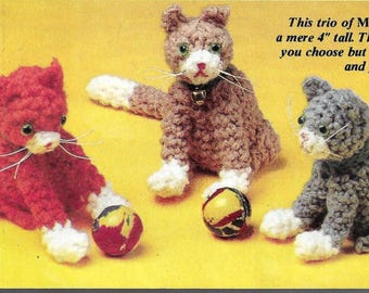 May include: Three crocheted cat toys, each 4 inches tall, sitting up with their paws and tail tips in white. The cats are red, grey, and brown. The cats are playing with small balls.