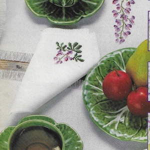 May include: Two green leaf-shaped plates with teacups and lemon slices. Each plate has a white napkin with a purple flower embroidery. A third green plate with fruit and a white napkin with a green flower embroidery.