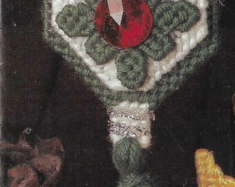 May include: A green and white crocheted ornament with a red jewel in the center. The ornament is hanging from a string and has a green tassel. The text "These easy-to-make treasures make great gifts all year long." is printed below the ornament.