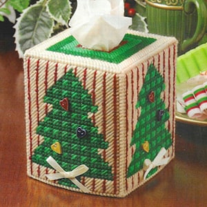 May include: A white canvas tissue box cover with a red and white striped pattern. The cover features two green Christmas trees with red and yellow buttons. The tissue box cover is handmade.