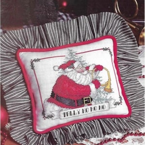 Peut inclure: Un oreiller blanc avec une bordure rouge et un volant à rayures noires et blanches. L'oreiller présente un motif de point de croix représentant le Père Noël jouant de la trompette avec le texte "Tally Ho Ho Ho".
