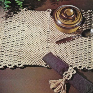 Könnte beinhalten: Ein beigefarbenes Makramee-Platzset mit Fransen an den Rändern und einem passenden Serviettenring. Das Platzset ist mit einem Knotenmuster gewebt und hat einen gewellten Rand. Der Serviettenring ist aus einem dunklen Stoff mit einer Quaste gefertigt.