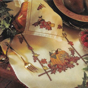 May include: A white embroidered table runner with an autumnal theme. The runner features a pumpkin, leaves, and berries in shades of orange, red, and brown. The runner is ideal for adding a touch of autumn to your table.