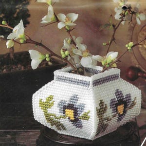 Könnte beinhalten: Eine weiße, geometrische Vase mit floralem Design, die Zweige mit weißen Blüten hält. Die Vase besteht aus einem strukturierten Material und weist ein lila-gelbes Blumenmuster auf. Die Vase steht auf einem dunklen Holzständer.