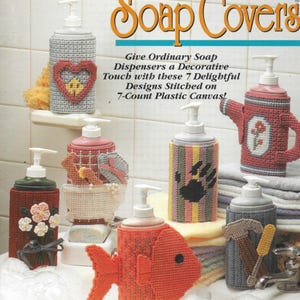 Op de afbeelding: Zeven zeepdispenserhoezen van plastic canvas met verschillende ontwerpen, waaronder een hart, een vis, een berenpoot, een vogel in bad, een gieter, een hamer en een zaag, en een boeket bloemen. De tekst "Soap Covers" staat bovenaan de afbeelding. De tekst "Give Ordinary Soap Dispensers a Decorative Touch with these 7 Delightful Designs Stitched on 7-Count Plastic Canvas!" staat onder de titel.