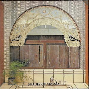 Op de afbeelding: Een gehäkelde macramé-gordijn met een franje, in crèmekleur, hangt boven een raam met houten jaloezieën. De tekst "Shades of the Sea" is onder het raam gedrukt.