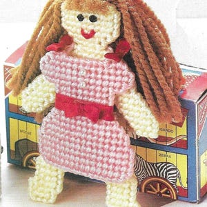 May include: A crocheted doll with brown yarn hair, a pink and white gingham dress, and a red bow. The doll is standing on a yellow and brown background.