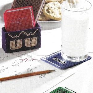 May include: A purple plastic container with a white and brown design holds three plastic canvas coasters. The coasters are red, brown, and blue with white lettering. The coasters say "Post It", "Relax", and "Pennies". A glass of water and a pencil are on a white surface with handwritten notes.