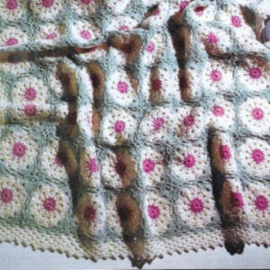May include: A crocheted baby blanket with a white and green floral pattern. The blanket has a crocheted border and is folded in half.