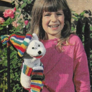 May include: A young girl wearing a pink sweater holds a white crocheted stuffed animal wearing a colorful striped scarf. The stuffed animal has a black nose and black eyes.