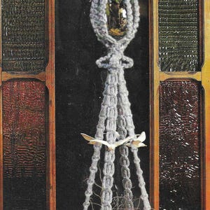 May include: A grey macrame plant hanger with a tassel hanging from the bottom. The hanger is in front of a stained glass window with the text "SEASCAPE Pattern on Page 12".