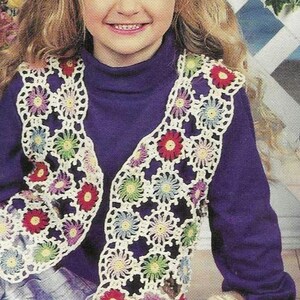 Peut inclure: Une jeune fille portant un pull à col roulé violet et un gilet crocheté avec des fleurs colorées. Le gilet est fait de fil blanc et a un bord festonné. La fille est assise sur un sol en bois avec une poupée portant un gilet similaire.