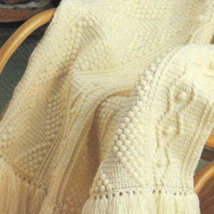 May include: Cream-colored knitted blanket with a textured pattern and fringe. The blanket features a combination of cable knit and bobble stitch designs. The blanket is draped over a wooden chair.