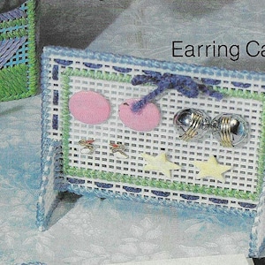 May include: A handmade earring caddy with a white grid design, blue and green trim, and the text "Earring Caddy". It features pink circles, silver earrings, and star-shaped accents. A matching bag is in the background.