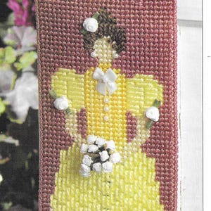 Pode incluir: Um pendurado de parede de ponto de agulha de tela plástica amarelo e branco com uma mulher em um vestido amarelo com flores brancas e um laço branco. A mulher está segurando um buquê de flores brancas. O pendurado de parede é emoldurado com uma borda bordô e tem uma guarnição de renda branca.