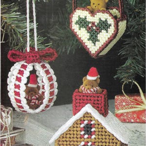 May include: Three handmade Christmas ornaments featuring a gingerbread house, a heart-shaped basket with a teddy bear, and a round ornament with a teddy bear inside. The ornaments are made with plastic canvas and yarn.