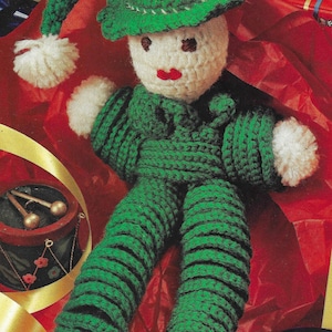 May include: A green crocheted elf doll with a white pom-pom on its hat. The doll is wearing a green and white striped outfit and has a white face with red lips. The doll is sitting on a red and white patterned surface.