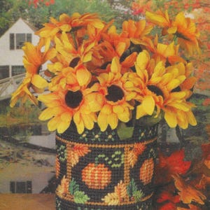 May include: A decorative vase filled with vibrant yellow and orange sunflowers. The vase is black with a cross-stitch design featuring pumpkins and fall leaves. The background shows a blurred landscape with a house and autumn foliage.