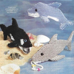 May include: Three plastic canvas sea creatures: a blue dolphin, a black and white orca, and a gray shark. The orca is holding a gold coin in its mouth and the shark is holding a string of colorful beads.