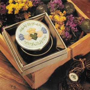 May include: A small round box with a green, yellow and blue Celtic knot design on the lid. The box is sitting in a wooden box with dried flowers. A gold chain with a matching pendant is hanging from the wooden box.