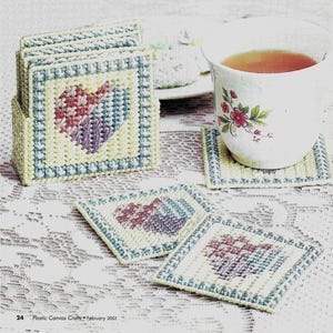 May include: A set of square, handmade coasters with a heart design in shades of red, purple, and blue. The coasters are in a holder, with a teacup on one. The coasters are yellow with a blue border.