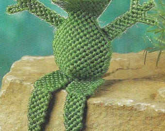 May include: A handmade green crochet frog with white accents, sitting on a rock. The frog has outstretched arms and legs, with a red mouth and white markings on its eyes. The frog is a whimsical decorative item.