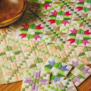 May include: Four crocheted placemats and four coasters in a geometric pattern with green, pink, white, and beige yarn. The placemats and coasters are arranged on a wooden surface.